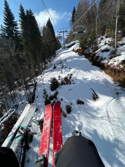 The image is a first-person perspective taken from a ski lift, overlooking a snowy ski slope near the city of Stoikite, Bulgaria. Below are tracks in the snow and patches of grass are visible. A mix of deciduous and coniferous trees populate the mountainside. Four pairs of skis are visible, two red and two blue/gray, hanging from the ski lift and belonging to the occupants. The ski lift structure, including the cable and chairs, is visible in the upper left of the image. The sky is visible on the left side of the image with a few white clouds.