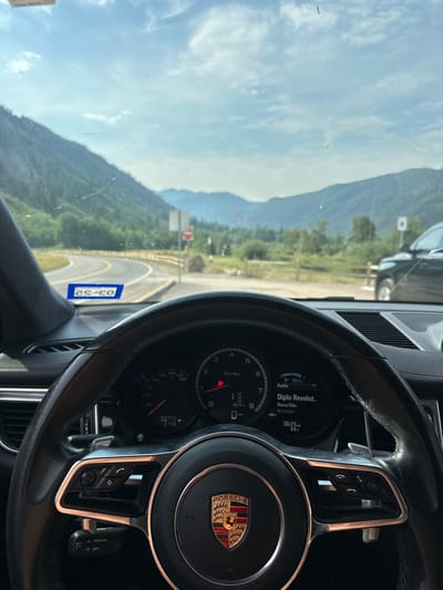 An image captured from the driver's perspective inside a stationary Porsche vehicle, identified by its steering wheel and dashboard, reveals a mountainous outdoor scene. The vehicle's dashboard shows an odometer reading of 59,777 miles, a speedometer at 0 MPH, and a digital display indicating "Diplo Revolution" audio playing, with the date 08/14/2023 and time 08:23 AM. The windshield exhibits smudges and minor cracks, and a reversed reflection of a license plate (reading "23.30") is visible on the lower left.
Outside the vehicle, a winding asphalt road with double yellow lines curves into the midground, flanked by a wooden guardrail and abundant green vegetation. To the right of the road, a white square sign with a red circular "No Entry" symbol stands on a pole. A large natural rock formation is situated behind this sign. Further right, a black SUV is parked in a pull-off area.
In the background, under a partly cloudy blue sky, a series of large, tree-covered mountains stretch across the horizon. A single red paraglider is visible airborne in the distant sky, between two mountain peaks. This scene is located in the mountainous region surrounding Aspen, United States, consistent with popular paragliding launch and landing areas near the city's scenic mountain roads, such as Maroon Creek Road or Castle Creek Road.