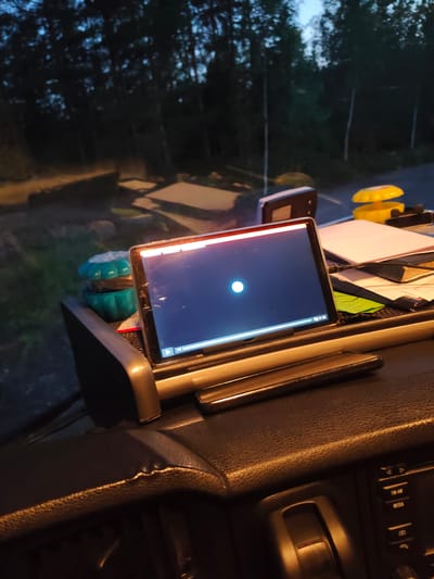 The image depicts the interior of a vehicle, possibly a truck, at dusk or night. A tablet computer is mounted on the dashboard, displaying a white circular icon on a dark blue screen. To the left of the tablet, a turquoise, multi-tiered object, possibly an air freshener or a decorative item, is visible. Further to the right, a stack of papers and a yellow object, resembling a hazard warning light or a cap, are present. The background shows a dimly lit forest area with trees. The vehicle's dashboard is visible in the foreground, with a textured, dark surface and some buttons of the audio system. The scene does not contain any people or specific landmarks that would indicate a location in Lohja, Finland, or any other city.