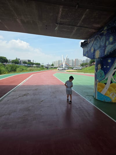 A young boy in a light blue t-shirt and grey shorts walks on a red track beneath a concrete overpass. The overpass is adorned with a mural depicting Vincent van Gogh's "Starry Night" and a portrait of Van Gogh himself. The track features a green section to the right of the red path, separated by a white line. In the background, several cars are parked, and a modern bridge with colorful arches spans a waterway. The cityscape features numerous apartment buildings and some trees. The sky is clear with a few scattered clouds. The scene appears to be part of an urban park or recreational area.