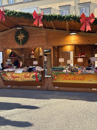 A detailed view of a festive outdoor market stall, likely part of a Christmas market, located in a city, specifically identified as Lucca, Italy. The wooden stall structure features two adjacent sections, each with an open counter display and an overhead roof. The roofline is adorned with evergreen garlands, small lights, and multiple large, glittery red bows. A prominent Christmas wreath with a large golden star is centered on the upper left section of the stall.

The left stall counter displays folded textiles, including sweaters and scarves, under a panel decorated with "Joyeux Noël" in red script against a golden, sparkling background with a red ribbon and ornaments. Behind this counter, a partially visible individual, wearing a dark jacket and a red scarf, attends to items on shelves displaying small bottles, decorative figurines, and price tags. A tall mirror reflects the stall's interior and a blurred view of an area beyond.

The right stall counter exhibits numerous folded "Foulards" (scarves) in various patterns and colors, situated below a panel with "Foulards" in red script against a similar festive golden background with red ornaments. Behind this counter, shelves hold wrapped gifts, a small Christmas tree, a red Santa hat, and additional price tags.

The ground in the foreground is a light-colored, textured pavement with distinct, elongated shadows stretching from the left, indicating direct sunlight. In the background, above the stall's roof, a light-colored stucco building with multiple windows, framed by decorative sills and equipped with light-colored shutters, is visible under a clear sky. The scene is bright, suggesting daytime conditions.