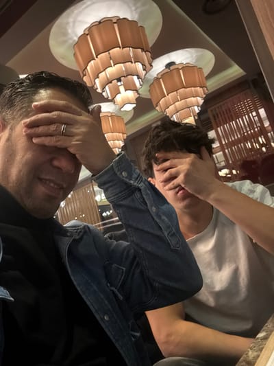 Two people cover their faces with their hands, appearing playful. The person in the foreground is wearing a denim jacket over a black shirt, smiling slightly. A silver ring is visible on their left hand. The person in the background wears a white shirt. The setting seems to be indoors with distinctive pendant lights illuminating the scene. The image is rotated slightly, adding to the casual feel.