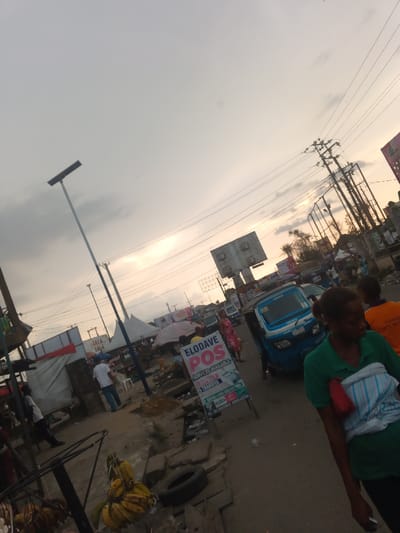 A street scene with a cloudy sky overhead. Power lines stretch across. People are walking, and a blue vehicle is visible. A sign advertises "ELODAVE POS SERVICES AVAILABLE". Bunches of bananas hang from a stand. The ground is rough and littered.