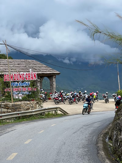 The image depicts a scene at a roadside establishment, likely a rest stop or cafe, in Lùng Tám Commune, Vietnam. The establishment features a rustic wooden roof and stone construction, with a prominent sign bearing Vietnamese text and the words "Vip Coffee" and "Coffee & Drinks." A large collection of motorcycles is parked in the foreground and to the side of the building, suggesting a popular stopping point for travelers. Several individuals are visible, either on or near the motorcycles, with one person operating a light blue motorcycle and moving down the paved road. The road curves to the right and is bordered by a concrete barrier and a rocky embankment. In the background, a vast expanse of mountains stretches into the distance, shrouded in dramatic, cloudy skies, indicating a potentially remote or mountainous region. The overall atmosphere suggests a stopover point on a scenic route, catering to motorcycle enthusiasts.