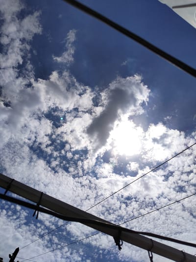 This is a low-angle, full-shot photograph of a bright, sunny sky filled with scattered white clouds. The sun is partially obscured by clouds, creating a bright halo effect. In the foreground, there are several dark, horizontal power lines and a section of what appears to be electrical conduit. The image is taken outdoors, likely in an urban environment given the presence of electrical infrastructure. There are no people or animals visible in the frame. The scene is consistent with a daytime view of the sky from ground level, looking upwards.