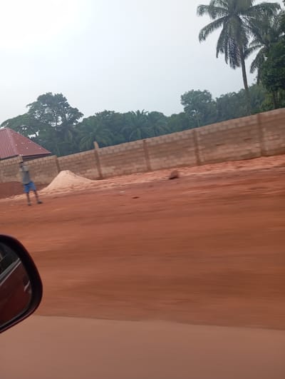 In Imilike, Nigeria, an overcast sky looms over a construction site. A person, wearing blue shorts and a grey t-shirt, stands near a pile of sand, their posture suggesting they are engaged in some activity. To the left, a building with a red roof is partially visible. A low brick wall runs horizontally across the scene, bordering the construction area. Palm trees and other dense foliage are visible in the background, indicating a tropical environment. The foreground is dominated by a dirt road, appearing reddish-brown, with motion blur suggesting the image was taken from a moving vehicle. A car's side mirror is visible in the bottom left corner, further confirming the perspective.