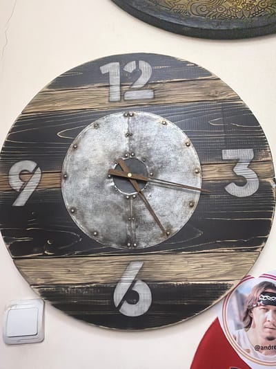 A wooden clock with a metal face and Roman numerals is mounted on a textured wall. The clock has dark wood planks with lighter wood accents. The numbers 12, 9, 3, and 6 are visible. The clock hands are made of dark wood. A white light switch is visible on the wall to the left of the clock. A circular item with a picture of a man is in the lower right corner, partially overlapping the clock. The man in the photo has a bandana on his forehead with a star design and is wearing a white t-shirt. The text "@andre" is visible below the man's image. The scene appears to be indoors. The location is identified as Maloyaroslavets, Russia.