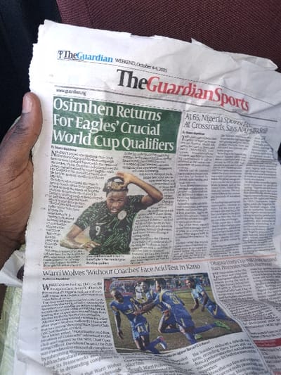 A crumpled, off-white copy of "The Guardian Sports" newspaper, dated "WEEKEND, October 4-5, 2025", is held by a dark-skinned hand on the left side of the frame. The newspaper's primary headline, highlighted in green, reads "Osimhen Returns For Eagles' Crucial World Cup Qualifiers". Below this, a photograph depicts a male soccer player, likely Victor Osimhen, wearing a dark green Nigeria national team jersey with the number "9" visible, his hands near his head. To the bottom-center, an article titled "Warri Wolves 'Without Coaches' Face Acid Test In Kano" accompanies a photograph of multiple male soccer players in blue and yellow uniforms actively engaged in a match on a green field. The background consists of a blurred, reddish-brown surface, possibly upholstery. This scene is observed within the city of Kungana, Nigeria.