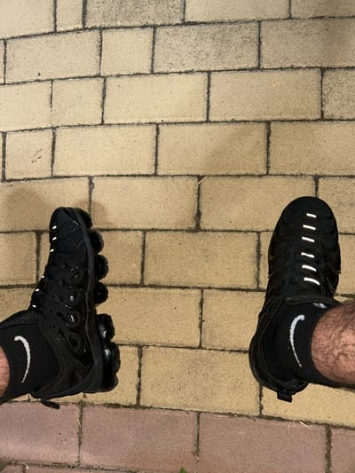 A top-down photographic view depicts two human feet, clad in black socks and black Nike Vapormax Plus sneakers, resting on a light-colored, rectangular brick paved surface in the city of Dzhebel, Bulgaria. The paving comprises individual bricks arranged in a consistent pattern with dark grey grout lines. Visible portions of the lower legs exhibit hair. Both feet are adorned with black ankle socks featuring a white Nike swoosh logo positioned on the outer side of each ankle. The left sneaker is oriented to reveal its distinctive side profile, including the segmented clear sole and a textured upper. The right sneaker is viewed from directly above, showcasing several parallel, reflective white strips across the instep of the shoe. The scene is illuminated by an ambient light source, accentuating the reflective elements on the footwear.