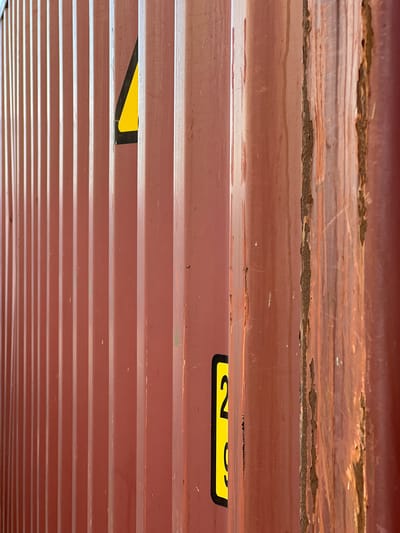 The image shows a close-up, abstract view of the corrugated metal exterior of a shipping container. The perspective is looking upwards, with multiple horizontal ridges of the container dominating the frame. The metal is painted a reddish-brown color, showing signs of wear and tear, including rust patches, scratches, and some dirt.
In the upper part of the image, there are more significant areas of rust and flaking paint, revealing the underlying metal and texture.
Two yellow warning labels with black text and borders are visible. One label, partially visible on the right side, appears to have the numeral "2" and other text. Another yellow triangle with a black border is visible in the lower-middle section of the image.
The overall impression is one of industrial decay and the passage of time. There are no people or clear indications of a specific event. The lighting suggests it is daytime, but no specific time or weather conditions are discernible. The location context provided is Újezd u Brna, Czechia, which, combined with the shipping container, suggests an outdoor industrial or transportation-related setting.