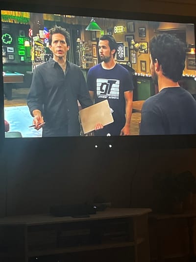 A scene from "It's Always Sunny in Philadelphia" is displayed on a television. Charlie Kelly, wearing a black shirt, stands with a pen in hand. Dennis Reynolds holds a clipboard, sporting a blue shirt with "Japanese Automotive" printed on it. The background includes a dartboard, various pictures, and bar decorations, creating a lively and cluttered setting within Paddy's Pub.