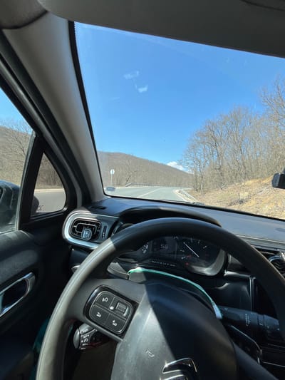 The image presents a forward-facing view from inside a vehicle, likely a Citroën, traveling on an asphalt road in a rural, hilly area. The location is specified as Shivarovo, Bulgaria.
The interior of the vehicle occupies the lower half of the frame, showcasing the top portion of a dark-colored steering wheel with integrated control buttons on its left spoke. Behind the steering wheel, the instrument cluster is partially visible, displaying the number "56," presumed to be the vehicle's speed, along with several illuminated indicator lights. An air vent on the far left of the dashboard features a circular air freshener attached. Minor reflections are present on the windshield and dashboard.
The exterior view, visible through the windshield, shows a two-lane asphalt road curving to the right in the mid-distance. A metal guardrail runs along the right edge of the road. On the left side of the road, a circular white road sign with a red border is visible. The surrounding landscape consists of rolling hills covered with bare deciduous trees, indicating a season devoid of full foliage, possibly late winter or early spring. A distant, lighter-colored peak or patch is discernible on the horizon to the right of the road. The sky above is clear and blue. No individuals are present in the observable scene.