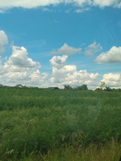 A wide shot of a rural landscape in Colonița, Moldova, captures a vast expanse of green grass under a vibrant blue sky filled with fluffy white clouds. Several scattered houses are visible in the distance, nestled amongst trees. The image appears to have been taken from a moving vehicle, suggesting a journey through the countryside. The overall impression is one of peaceful, natural beauty on a sunny day.