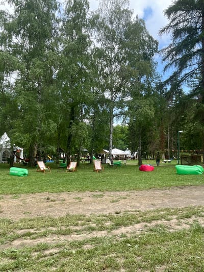 This image captures an outdoor recreational area within Sremski Karlovci Municipality, Serbia, on a partly cloudy day. The scene is dominated by numerous trees, primarily birches with light-colored bark and dense green foliage, interspersed with darker conifers on the right side, framing a bright blue sky with white clouds. The ground consists of a grassy lawn showing significant wear with patches of exposed dirt and discernible tracks in the foreground and midground.
In the immediate foreground, a bright green inflatable lounge bag marked "PLAYRIX FEST" is positioned on the left. To its right, three wooden-framed deck chairs with light-colored canvas seats are arranged around a small, dark, round table. Further right, a red inflatable lounge bag and another green inflatable lounge bag are visible on the grass.
In the midground and background, several white temporary tent structures are set up, indicating an event or gathering. On the far left, a white peaked tent with clear window panels is visible, near which a red hammock is strung between two trees. Further back, larger white marquee tents are present, with indistinct figures of people moving around them, suggesting activity. A yellow slide is faintly visible in the deeper background, near the center. On the right side, a person wearing a dark top and lighter trousers is walking away from the viewer, towards the right. A string of colorful triangular pennant banners (orange, green, red) is strung between trees and a light pole on the far right, adding a festive element to the scene. The overall impression is that of a temporary outdoor event in a natural park-like setting.