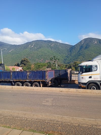 The image captures a sunlit road scene in Namanga, Kenya, featuring large trucks against a backdrop of mountains. In the foreground, a paved road with a rough curb stretches across the lower portion of the frame.
Two heavy-duty trucks dominate the midground. On the right, the front section of a white semi-truck cab is visible, facing left. The cab features a black and white logo with "K&V" prominently displayed, beneath which the text "EXCELLENCE IN LOGISTICS" is visible, followed by smaller, illegible lines of text. To its left, a long blue flatbed semi-trailer with high, slatted sides is parked or moving slowly. This trailer has multiple axles and wheels visible underneath, designed for hauling goods. Another dark-colored truck or trailer is partially visible behind the blue one, suggesting multiple vehicles.
In the background, a striking range of large, green mountains, densely covered in trees, extends across the horizon. Scattered white clouds punctuate the otherwise clear, bright blue sky above the mountains. At the base of the mountains, a few buildings with reddish-orange roofs are faintly visible amidst the foliage. The scene is illuminated by bright daylight, suggesting it is morning or early afternoon, with clear and sunny weather. The trucks appear to be either parked or moving slowly on the side of the road, indicating a transport route through a mountainous region.