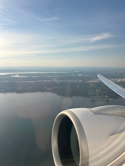 The image, taken from an airplane window, shows a view of the city of Notre-Dame-de-l'Île-Perrot, Canada, from above. In the foreground, the engine of the airplane is visible, with its metallic casing and a glimpse of the propeller blades within. The airplane's wing extends towards the right side of the frame. Below, a large body of water, likely Lake Saint-Louis, stretches across the landscape. Beyond the water, the urban sprawl of Notre-Dame-de-l'Île-Perrot unfolds, characterized by a dense network of buildings, trees, and roads. The sky above is a clear blue, with a few wispy clouds, suggesting it is a bright, possibly late afternoon, day. The overall scene depicts a typical aerial view of a Canadian city situated near a significant body of water.