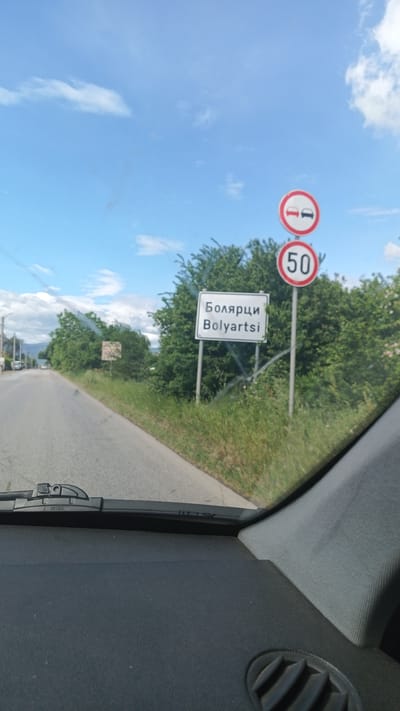 The image depicts a view from inside a vehicle, likely a car, driving on a road toward the village of Boliartsi, Bulgaria. A sign displaying "Болярци Boliartsi" marks the entrance to the village. Above the village sign, there are two road signs: one indicating "Overtaking Prohibited" and another showing a speed limit of 50 km/h. The road is asphalt, with green vegetation growing on the side. The sky is blue with scattered clouds. The interior of the vehicle, including the dashboard and windshield, is visible.