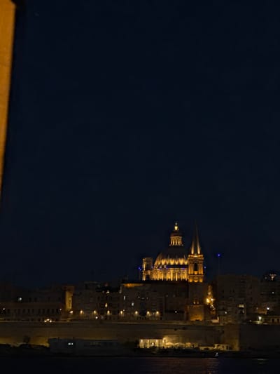 The image depicts a nighttime scene of a coastal city. The primary subjects are a large, illuminated dome and spire structure, likely a cathedral or major landmark, with smaller buildings clustered around it. The architecture suggests an older European style, with many windows lit from within, indicating residential or commercial activity. The buildings are built along a waterfront, with the dark water of the sea or harbor visible on the right side of the frame.
The time of day is clearly night, as evidenced by the dark sky and artificial lighting illuminating the buildings. The weather appears calm, with no visible signs of rain or strong winds. The scene is likely set in a city with a distinct architectural heritage, and the presence of numerous lights suggests it is a populated area. There are no people visible in the image.
Notable details include the warm, golden illumination of the main structure, contrasting with the cooler blue lights visible on some of the surrounding buildings and the spire. The overall mood is one of quiet urban life at night, with the illuminated buildings creating a picturesque view against the dark expanse of the sky and sea. The image is oriented vertically, with the buildings appearing to be on the right side of the frame. While there is no visible text, the architecture and lighting strongly suggest a European city. The mention of Gżira, Malta, provides specific location context, and the prominent dome and spire could be recognizable landmarks within that area, possibly St. John's Co-Cathedral or a similar significant building.