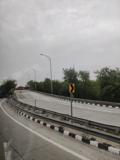 The image shows a highway curve on a cloudy day in Unknown, India. The road is asphalt with white lane markings and a black and white striped barrier along the side. There are several streetlights visible along the road, and a traffic sign indicating a curve to the right. Trees and foliage line the side of the highway. A red truck is visible in the distance. The perspective suggests the photo was taken from a vehicle.