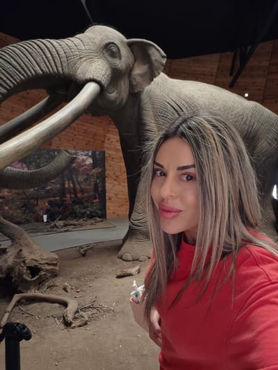 A woman, with long blonde and brown streaked hair, wearing a red long-sleeved shirt, is taking a selfie in front of a large, realistic model of an extinct elephant, likely a mammoth, within an indoor exhibit. The elephant model is positioned centrally in the background, with its tusks prominently displayed. The environment appears to be a museum or natural history exhibit, indicated by the earthen floor, wooden paneled walls, and a backdrop depicting a forest scene with other extinct creatures. Some broken tree branches and logs are scattered on the ground in the foreground. The lighting is dim, creating a slightly shadowed atmosphere. The woman is smiling and looking directly at the camera. The location is specified as the Dorkovo paleontological site in Bulgaria.