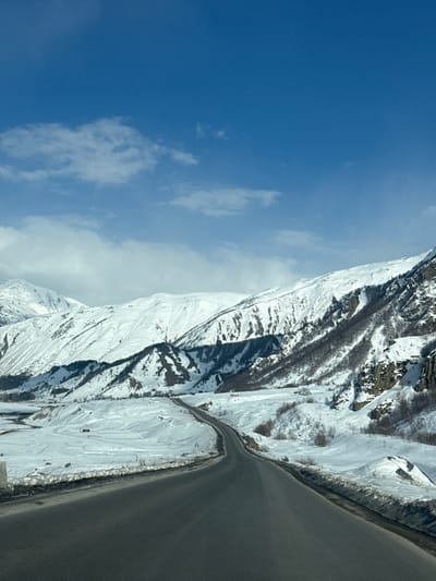 The image displays a winter mountainous landscape in Sioni, Georgia. A two-lane asphalt road, clear of snow, extends from the foreground and curves right into the middle ground, then straightens before disappearing into the distant valley. The ground surrounding the road is covered in a substantial layer of white snow, forming undulating terrain and slopes.

Snow-capped mountains dominate the background and midground, featuring jagged peaks and exposed dark rock faces and ridges on their lower and upper slopes. Patches of bare deciduous trees are visible on some mountain slopes and within the valley, appearing as dark, dense clusters against the white snow. The sky is bright blue with scattered white cumulus clouds, occupying approximately the upper third of the frame. A small concrete or stone road barrier is visible on the left side of the foreground, adjacent to the road. No individuals, vehicles, or other significant man-made structures are present, indicating a natural, remote setting under clear winter conditions.