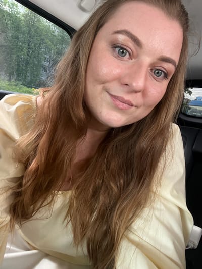 A selfie of a Caucasian woman with long brown hair, light skin, and blue eyes, wearing a light yellow blouse, is taken inside a vehicle. The setting appears to be daytime, judging by the lighting. Visible through the car window are green trees and a building, possibly indicating a residential area. The location is not identifiable as Balabanovo, Russia based on the visible information. The woman is looking directly at the camera with a slight smile. A white object and dark panel are visible in the background within the car.