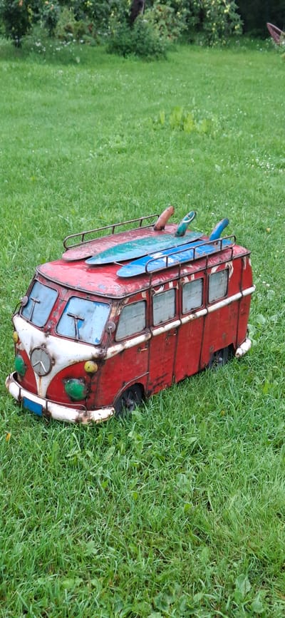 A vintage-style, red and white metal camper van replica, adorned with three surfboards on its roof rack, sits in a grassy outdoor area. The replica van has a classic Volkswagen Type 2 silhouette, complete with round headlights and a prominent front grille. The surfboards are colored blue and green, with one partially obscured by another. The van is resting on a patch of green grass, with some scattered dandelions visible. The background consists of more green grass and blurred foliage, suggesting a natural outdoor setting. The image is taken from a slightly elevated angle, looking down at the van.