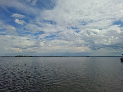 The image captures a wide expanse of dark, rippling water under a dramatic sky filled with a mix of white and grey clouds. In the distance, a thin strip of land is visible, featuring a cluster of green trees and a faint outline of buildings and industrial structures, possibly hinting at the presence of the city of Verhnij Uslon, Russia. The horizon line is clearly defined, separating the water from the sky. On the far right of the frame, a portion of a wooden structure, possibly a pier or a boat dock, is visible, suggesting a waterfront location. The overall atmosphere is overcast and somewhat moody.
