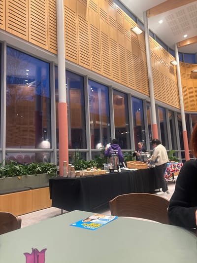 The image depicts an indoor event space within the Hub building at Edge Hill University in Ormskirk, West Lancashire, United Kingdom. The scene shows a modern, well-lit area with high ceilings, featuring light-colored wooden slatted walls and a long expanse of floor-to-ceiling windows. Outside the windows, the environment is dark, indicating evening. Along the window base is a planter box filled with green foliage.

In the mid-ground, a black-draped table serves as a refreshment station. Three individuals are present at the table. On the left, a person wearing a purple "Campus Connector" hoodie faces away from the camera, attending to a silver beverage dispenser. In the center, a woman with light hair and a lanyard stands facing right. On the right, a woman in a light-colored cardigan and dark trousers selects items from a wicker basket on the table. The table holds stacks of brown paper cups, various food items in silver foil trays, and yellow packets. To the far right of the table, a banner featuring international flags is visible.

In the foreground, the edge of a light green table is visible, with a blue leaflet titled "FANCY WINNING £100?" placed upon it. A person with reddish-brown hair is partially visible, seated at this table on the right. The room features multiple slender white columns with peach-colored bases, and recessed lighting in the ceiling.