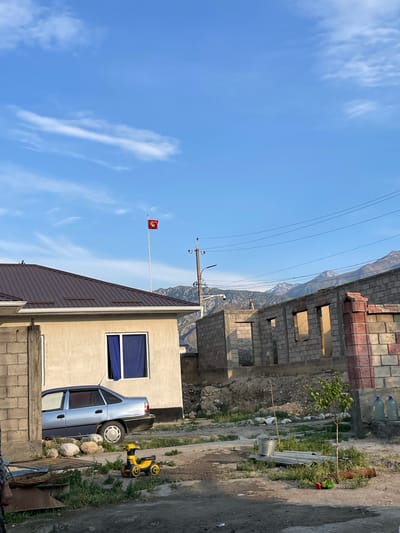 The image depicts a street scene in Ak-Sai, Kyrgyzstan. A light blue sedan is parked in front of a beige building with a dark brown metal roof. To the right of the building, the skeletal remains of a brick structure under construction are visible. In the background, majestic mountains form a backdrop under a clear blue sky with scattered clouds. A flag with a yellow sun and a tündük (a yurt crown) on a red field, representing Kyrgyzstan, flies from a tall flagpole. A yellow toy ride-on car is positioned on the ground in front of the parked car. The overall impression is one of a developing residential area with a clear view of the natural landscape.