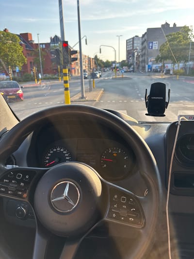 The image is taken from the driver's perspective inside a Mercedes-Benz van, looking forward through the windshield. The van is stopped at a traffic light in the city of Boom, Belgium, at the intersection of the N16 and Boom's main street, located near the local church. The van's dashboard, including the steering wheel with the Mercedes-Benz logo and airbag indicator, and the instrument cluster displaying speed and engine RPM, are visible. A Grundig phone holder is attached to the windshield. Outside, typical urban scenery of Boom is depicted, with buildings lining the street, trees, streetlights, and other vehicles. The sun is low in the sky, casting long shadows and creating a warm, golden light, suggesting it is either early morning or late afternoon. The traffic light is red, indicating the van is temporarily halted.