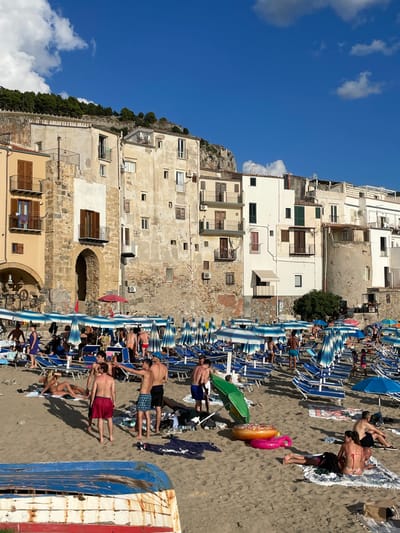 The image displays a busy beach scene in Cefalù, Italy, specifically at the base of the historic multi-story buildings fronting the coast, with the prominent Rocca di Cefalù rising in the background. The sky is clear blue with scattered white cumulus clouds, indicating bright daytime conditions.
In the lower-left foreground, a dilapidated, overturned wooden boat, painted in chipped white, blue, and red, rests on the sand. The mid-ground is dominated by a sandy beach densely populated with numerous blue and white striped beach umbrellas and corresponding blue sun loungers arranged in rows. Approximately 25-30 individuals are visible on the beach. Most are adults in swimwear, engaged in leisure activities such as sunbathing on loungers or towels, standing, and walking. A distinct group of five men is positioned mid-frame, one gesturing with an outstretched arm to the right. Two inflatable rings, one orange/pink and one yellow/pink, are on the sand, along with a green beach umbrella and various towels and clothing items.
The background consists of a continuous line of aged, multi-story buildings, varying in color from light yellow to off-white and exposed stone. These structures feature numerous windows with dark wooden shutters and multiple balconies with metal railings. An arched entryway is visible on the left-most building. Immediately behind these buildings, the steep, rugged rock face of the Rocca di Cefalù rises sharply, topped with dense green vegetation.