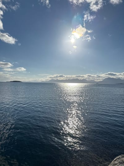 A seascape shot under a bright, sunny sky with a hint of clouds. The sun is positioned high and slightly to the right, casting a brilliant reflection across the calm water, creating a shimmering path of light. In the distance, a chain of mountains forms the horizon, partially obscured by clouds. To the left, a small island is visible. The foreground features the dark blue water, with a rocky shore on the right side of the frame. This view likely captures a scene along the coast of Harstad, Norway, given the characteristic landscape of fjords and mountains. No people or animals are present.
