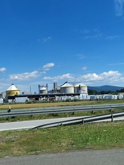 The image shows an industrial facility under a bright blue sky with scattered white clouds. The facility appears to be a biomass pellet plant, as suggested by the signage on one of the silos which reads "SCHWAIGER ROLL INDUSTRIE" and another with "GER PELLETS". There are large, conical silos, storage buildings, and stacks of bundled material, possibly wood pellets or lumber, outside. The plant is situated in a rural area, with a field of dry grass in the foreground and a wooded hillside in the background. A highway guardrail runs across the middle ground, separating the viewer from the industrial site. The image appears to have been taken from a moving vehicle on the highway. The location is identified as Hengersberg, Germany.