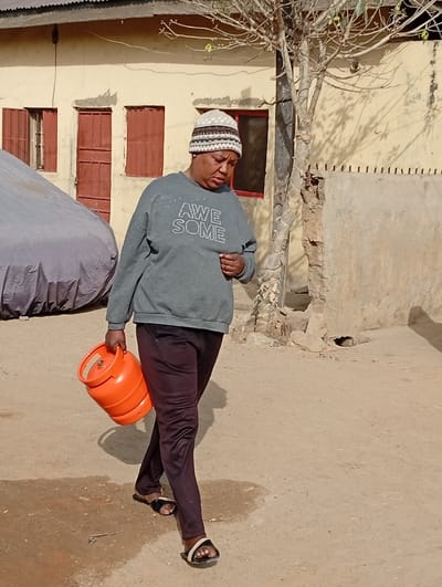 In Gayawa, Nigeria, a single individual walks rightward across a dry, dusty dirt path. The person wears a multi-patterned knit beanie, a grey long-sleeved sweatshirt with "AWE SOME" printed on the front, dark purple trousers, and black and white open-toed sandals. An orange gas cylinder is carried in the subject's right hand. The background features a light-yellow building with a flat roof, displaying multiple windows fitted with red shutters and frames; some shutters are open, others closed. A vehicle covered with a grey tarp is parked to the left of the building. To the right, a bare-branched tree stands, casting distinct shadows on a partially damaged wall section. The scene is outdoors under natural sunlight.