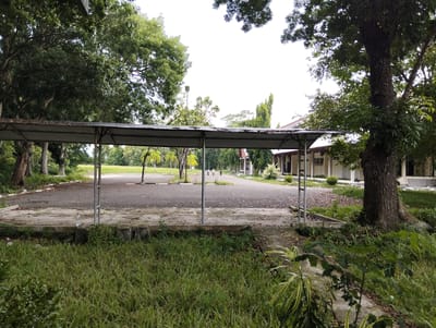 The image depicts an outdoor scene at an unidentified location in Palohgadeng, Indonesia. In the foreground, there is a covered walkway with a metal roof supported by pillars. The ground beneath the walkway is paved and appears weathered. Beyond the walkway, a paved open area, possibly a parking lot or courtyard, is visible, with some scattered leaves and debris. In the background, there is a building with a light-colored facade and a dark roof. Several trees are present, some with lush green foliage, and others are more sparse. The sky is overcast, contributing to the diffused lighting of the scene. There are no people clearly visible, though distant figures may be present in the background. The overall impression is one of an established, possibly underutilized, outdoor space within a larger complex.