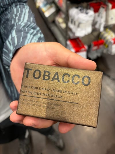 An overhead close-up shot captures a hand holding a rectangular box of "TOBACCO VEGETABLE SOAP." The hand, appearing to be an adult's, holds the box centrally against a soft, out-of-focus background. The person is wearing a grey and white patterned long-sleeved top or jacket.
The soap box is a warm, metallic gold or bronze color with a slightly speckled texture. Large, black, sans-serif letters spell out "TOBACCO" vertically along the left side of the box. Below and to the right of this, smaller black text is visible, reading: "VEGETABLE SOAP - MADE IN ITALY", "NET WEIGHT 250 G 8.75 OZ", "Made with vegetable ingredients.", and partially "leaves soft and velt" (likely "velvet").
The background is highly blurred, showing indistinct shapes and colors, primarily white, red, and grey, suggesting an indoor setting, possibly a store or a storage area in Luton, United Kingdom. The activity depicted is simply the act of holding and presenting the soap box, perhaps for inspection or photography. No specific time of day or weather conditions are discernible from the indoor context.