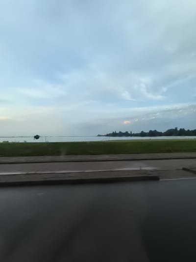 This image depicts a serene waterfront scene, likely taken from a moving vehicle, showcasing the tranquil waters of the Westeinderplassen in Aalsmeer, Netherlands. The foreground is dominated by a dark, reflective surface, possibly a road or pavement, followed by a strip of manicured green grass. Beyond the grass, the expansive Westeinderplassen stretches out, its surface calm and reflecting the overcast sky. A silhouetted object, possibly a small boat or structure, is visible on the water in the mid-ground, anchored by thin poles. The far bank of the lake is lined with a dense row of dark green trees, creating a natural silhouette against the horizon. The sky is a soft blue with diffused clouds, and a faint, warm-toned cloud formation is present in the distance, suggesting the possibility of a sunset or sunrise. The overall impression is one of quietude and natural beauty.