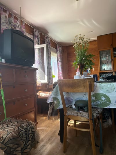 The image shows an interior view of a room, likely a living or dining area, featuring wooden walls and a wooden floor. The room is lit by natural light streaming in from a window and artificial light from ceiling fixtures.
Prominently in the foreground and center is a wooden dining table covered with a tablecloth featuring a green and white leaf pattern. A wooden chair with a woven seat is positioned at the table, with a green footstool on the seat. On the table, a vase of white flowers stands tall, surrounded by other objects including a box with Russian text, glasses, and a remote control.
To the left of the table is a dark wooden dresser with multiple drawers. A vintage television set sits atop the dresser, with an antenna extending upwards. A green string is attached to the side of the dresser, dangling towards the floor. A patterned fabric, possibly a rug or throw, is draped over a surface in front of the dresser.
The background reveals more of the room's features: a window with floral curtains, a cabinet with glass doors displaying various items, and more wooden paneling on the walls. The overall impression is of a lived-in, rustic interior, possibly a dacha or a rural home. The presence of the television and dining setup suggests a functional living space. The location is Yalguba, Russia.