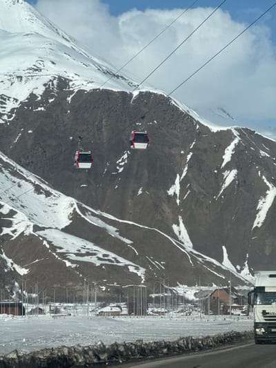 This image depicts a winter mountain landscape in Almasiani, Georgia, under a partly cloudy sky. The upper and mid-portions of the frame are dominated by rugged, snow-dusted mountainsides, featuring exposed dark rock formations interspersed with white snow patches and streaks. Two red and white gondola cabins are visible, suspended by cables and traversing horizontally across the mountain slope from left to right.
The lower section of the image shows a valley floor, heavily covered in white snow. Numerous tall, slender streetlights are arranged in parallel rows across this snowy expanse, suggesting a developed area. Several buildings, some with dark brown roofs, are scattered among the streetlights. In the immediate foreground, an asphalt road runs horizontally, bordered by a rough, discolored snow and slush barrier. On the right side of the road, the front portion of a white semi-truck, branded "IVECO" and displaying a partially visible Russian license plate, is visible, oriented towards the left. No identifiable individuals are present in the scene.