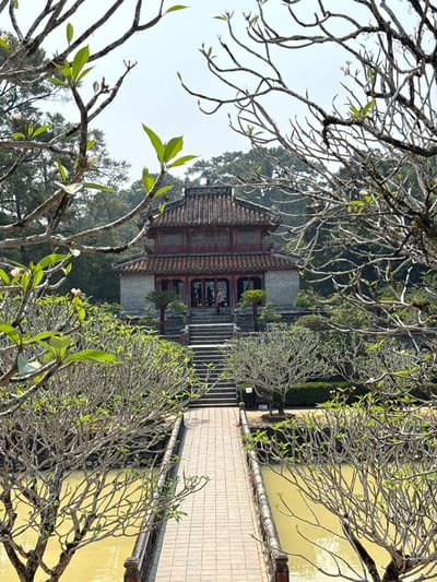 A landscape shot, taken at an angle, features a building with dark red accents and an intricate roof, set against a backdrop of lush, green trees and shrubbery, possibly located in the city of Kim Long Ward, Vietnam. In the foreground, leafless branches with some green leaves frame the view. A walkway extends from the bottom right towards the building, positioned off-center to the left. The walkway has a pond or similar body of water on either side. The sky is pale and overcast, suggesting the photo was taken during the day.