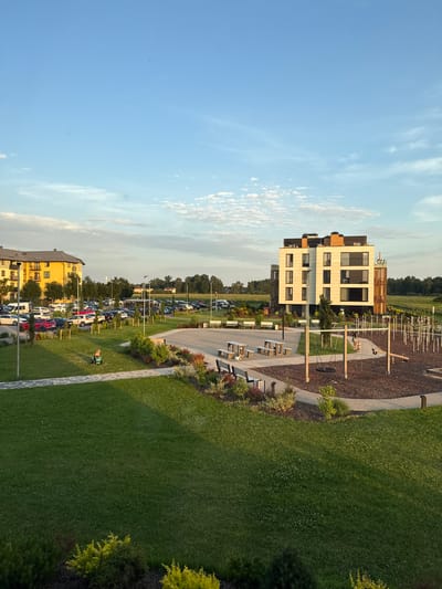 The image captures an outdoor scene in Mārupe, Latvia, likely a residential or communal area, bathed in the warm light of late afternoon or early evening. In the foreground, a meticulously maintained green lawn stretches across the frame, with a paved walkway winding through it. Beyond the lawn, a modern playground area is visible, featuring swings, climbing structures, and a soft mulched surface. Several benches are placed around the playground and along the walkways.

To the left, a bright yellow apartment building stands prominently, with numerous windows and balconies. A parking lot filled with various cars is situated in front of this building, bordered by rows of young trees. In the mid-ground, a contemporary, multi-story building with a light-colored facade and dark-framed windows is a focal point. Adjacent to this building is a wooden tower-like structure, possibly an architectural feature or part of the playground.

Further into the distance, a dense line of trees forms a natural boundary, and beyond that, open fields and sky are visible. The sky is a clear blue, with scattered wispy clouds. A single person, appearing to be a child, is seated on a small green ride-on toy on the lawn. Another person is seated on a swing in the playground. The overall impression is one of a well-developed and tranquil residential environment.
