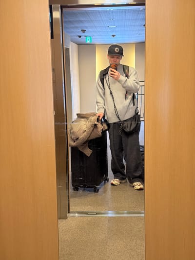 A man stands in front of a mirror, reflected with his luggage in an entryway or hallway. He wears a backwards baseball cap, grey sweater, dark pants, and sneakers. A small bag hangs at his side. He holds up a phone, taking a selfie. A dark suitcase with a light tan jacket sits on top, is to his left. The hall has a light carpet and textured wall paneling and ceiling. The exit sign is visible.