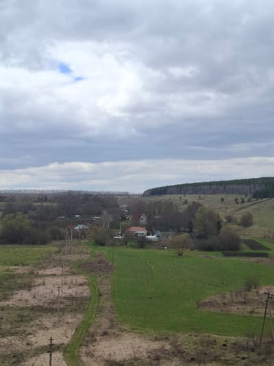 The image captures a broad rural landscape under a cloudy sky, likely in Yablonovo, Russia. The upper half of the frame is dominated by an overcast sky, filled with shades of white and grey clouds, with a small, distinct patch of blue sky visible towards the top left.

The lower half of the image presents a diverse terrain. On the right, expansive fields stretch out, featuring patches of vibrant green grass intermingled with areas of drier, brownish vegetation, possibly indicating early spring growth or varying land use. Several utility poles with power lines run horizontally across these fields.

Towards the center and left, a small village or hamlet is nestled amongst a cluster of trees, many of which appear to be deciduous and in an early stage of leafing out or still bare. Several houses with visible roofs are scattered within this wooded area. In the far distance, a dark, dense line of forest, likely coniferous, defines the ultimate horizon, contrasting with the lighter sky above.

The scene depicts a tranquil, unpopulated rural environment during daylight hours, with no specific activity taking place. There is no visible text in the image.