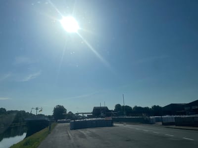 The image depicts a bright, sunny day in Edewecht, Germany. The sun is high in the sky, creating a strong glare and casting lens flares throughout the scene. The sky is a clear, bright blue with a few wispy clouds. In the foreground, there is an asphalt road with white lane markings. To the left of the road is a canal with calm water reflecting the sky and the surrounding greenery. A barge is docked on the canal, with two flags flying from its mast.

Beyond the canal and to the right of the road, there is a large industrial area. Piles of goods, likely wrapped in white plastic, are stacked high in rows. Buildings with dark roofs and walls are visible in the background, suggesting a commercial or industrial facility. Trees and other vegetation are scattered throughout the landscape, with a dense line of trees forming a backdrop to the industrial area.

The overall impression is one of a working industrial area on a clear, warm day. The presence of the canal and barge suggests that water transportation is a significant aspect of the location. The abundance of stacked goods indicates ongoing commercial activity. There is no visible text in the image that can be read. The time of day is clearly daytime, likely mid-morning or early afternoon given the sun's position and intensity. The weather is hot and sunny.