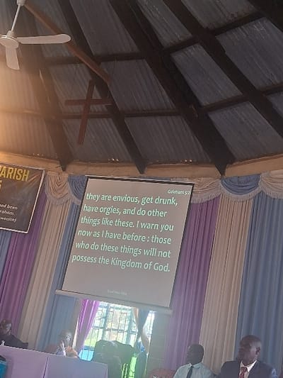 This image captures an indoor gathering in Muthambi ward, Kenya. A white projector screen displays biblical text from Galatians 5:21 of the Good News Bible, stating: "they are envious, get drunk, have orgies, and do other things like these. I warn you now as I have before: those who do these things will not possess the Kingdom of God." The backdrop features draped fabric in shades of light blue, purple, and white. Above, the ceiling consists of corrugated metal roofing supported by dark wooden rafters. A white ceiling fan and a suspended wooden cross are visible among the rafters. A window with metal grilles is visible through the drapes, showing greenery outside. In the foreground, four dark-skinned men are present. On the left, two men are seated behind a light-colored table; one wears glasses. On the right, two men are seated in the immediate foreground; one wears a light blue shirt and dark tie, the other a dark suit jacket and a red patterned tie. All visible individuals are seated and appear to be observing the projected content or participating in a meeting. A partial banner on the far left displays the word "PARISH."