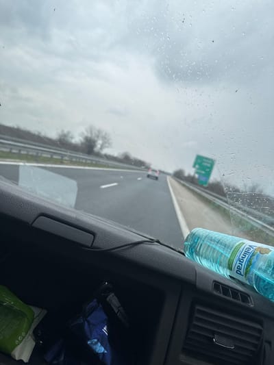 A forward-facing view from inside a moving vehicle on a multi-lane highway near Gabarnitsa, Bulgaria, under an overcast sky. The windshield is speckled with water droplets and streaks, suggesting recent precipitation. The highway ahead shows two lanes of travel in the same direction, marked by white dashed lines. A light-colored sedan with illuminated taillights is visible in the distance in the right lane. A blurred green highway sign is discernible on the right shoulder, beyond a metal guardrail. Bare trees line the highway in the background. The foreground displays the black, textured dashboard of the vehicle. On the passenger side, an open storage compartment contains a green and white packaged item on the left and several dark blue packaged items on the right. A clear plastic bottle of "VELINGRAD ACQUA" brand water rests horizontally on the dashboard to the right, next to air vents. A black cable runs across the dashboard's upper edge. No individuals are visible.