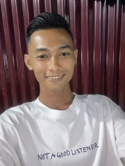 A young man is depicted in a close-up portrait, smiling broadly at the camera. He has short, dark, slicked-back hair and a cheerful expression, revealing his teeth. His skin has a slight shine, possibly from lighting or perspiration. He is wearing a plain white, long-sleeved t-shirt. Printed vertically on the front of his shirt, in dark, hand-written style capital letters, is the phrase "NOT A GOOD LISTENER". The background features a dark, reddish-brown corrugated surface, likely a metal wall or door, providing a textured backdrop with horizontal lines. The lighting appears direct, illuminating the man and his white shirt. The image captures a casual moment, with the subject conveying a friendly and approachable demeanor. While the specific location isn't visually evident, the provided context suggests Gampongaceh, Indonesia.