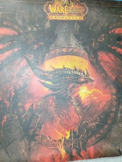 This media file is a photograph of a playmat featuring artwork from the game "World of Warcraft: Cataclysm." The artwork depicts a dragon-like creature with dark, scaly armor and glowing orange and yellow accents, seemingly in a fiery environment. The dragon's head is prominent, with sharp teeth and menacing eyes. The overall scene conveys a sense of power and destruction.
Visible text on the playmat includes the "World of Warcraft" logo and the word "Cataclysm" in a stylized font. There are also some decorative elements that appear to be runes or symbols around the central image.
The image itself is a photograph taken from a slightly elevated angle, with the playmat lying flat. The lighting in the photograph is uneven, and the texture of the playmat's fabric is visible. The photograph appears to have been taken indoors, with no specific environmental cues suggesting a particular time of day, weather, or location beyond the subject matter of the playmat itself. The artwork evokes a fantasy setting and a feeling of intense conflict.