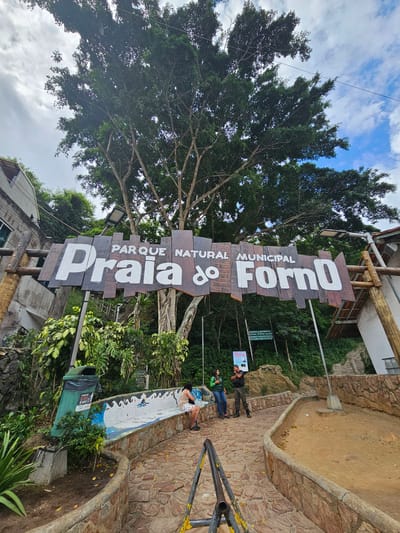 The image displays the entrance to the Parque Natural Municipal Praia do Forno in Arraial Do Cabo, Brazil, under a partially cloudy sky. A large, rustic wooden sign with white letters spelling "PARQUE NATURAL MUNICIPAL PRAIA do FORNO" forms an archway over a curved, reddish-brown cobblestone pathway. To the left of the path, a long, curved concrete bench with a blue and white wave mosaic design is integrated into a low stone wall. A green trash receptacle with a black liner and text "LUGAR DE LIXO É AQUI" stands beside the bench.
Three individuals are visible: one person is seated on the mosaic bench, wearing a light top and patterned shorts. Further along the pathway, near the entrance sign, two individuals stand. One, possibly a park ranger, is dressed in a dark uniform consisting of a green shirt, dark pants, and boots, interacting with another individual wearing a green top and blue jeans.
Dense green vegetation, including large trees with extensive canopies and thick undergrowth, covers the slopes surrounding the entrance. Parts of structures, including a grey-walled building on the left and a white-walled building with a red-tiled roof section on the right, are visible. Wooden steps ascend a hillside on the left side of the main sign. A green sign behind the main archway reads "PRESERVE A NATUREZA!", and a white sign with a bird icon states "NÃO ALIMENTE AS AVES" (Do not feed the birds). A yellow and black metal barrier is positioned in the immediate foreground. Overhead power lines are present against the sky.